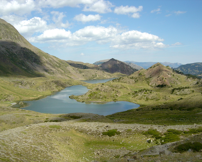 A high-altitude landscape with green slopes devoid of shrubs and a small lake reflecting the cloudy sky