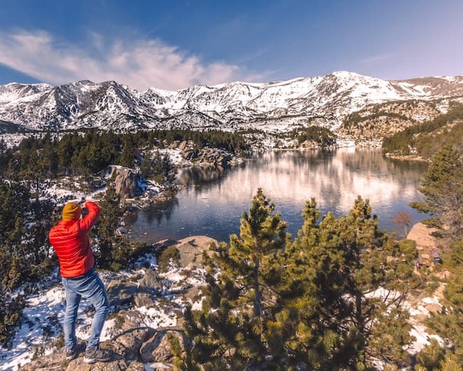 Un excursionista con una chaqueta roja fotografía un lago que refleja las cumbres nevadas.